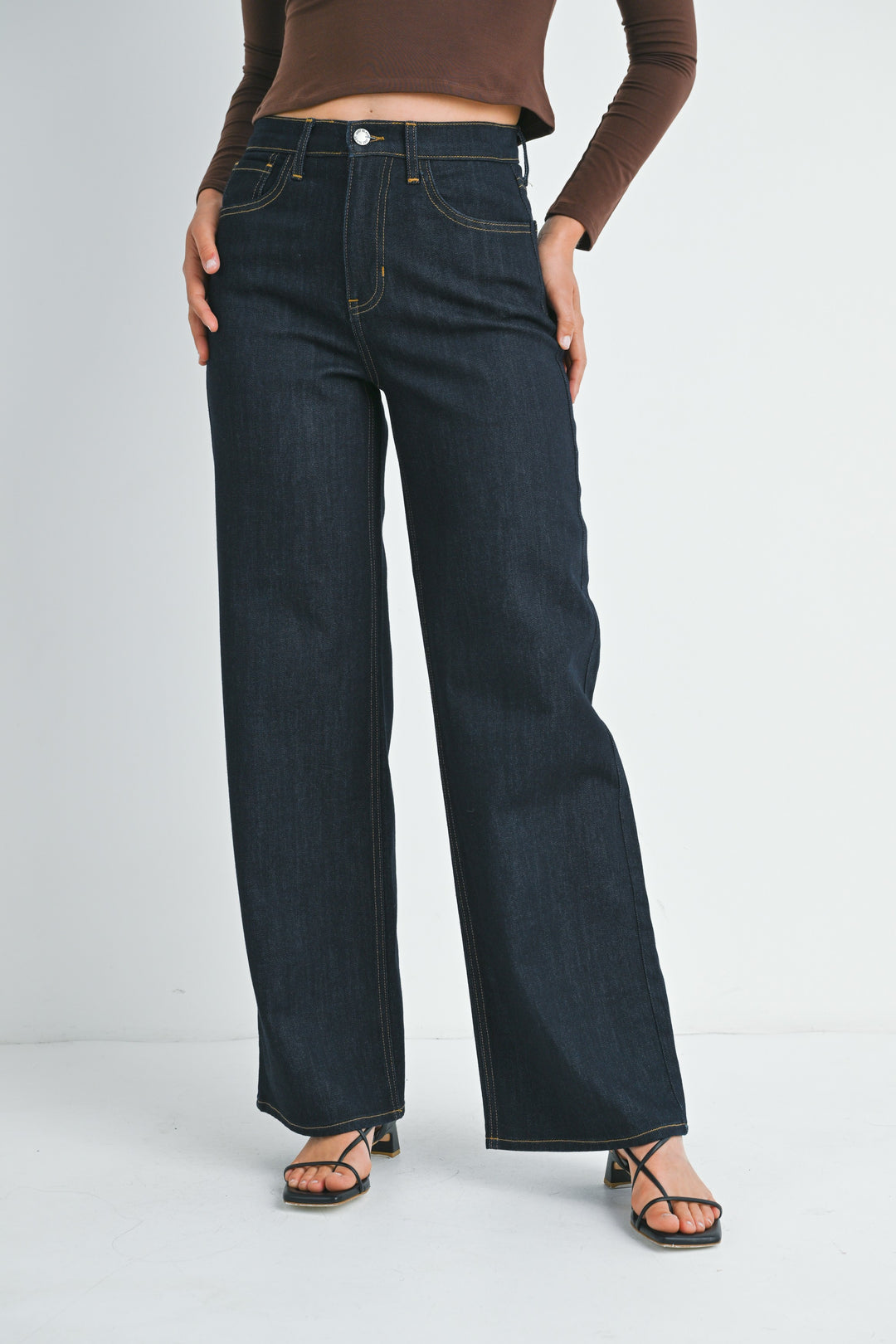 A model wearing dark wash wide-leg jeans, a brown long-sleeve crop top, and black strappy sandals, standing against a light gray studio background.