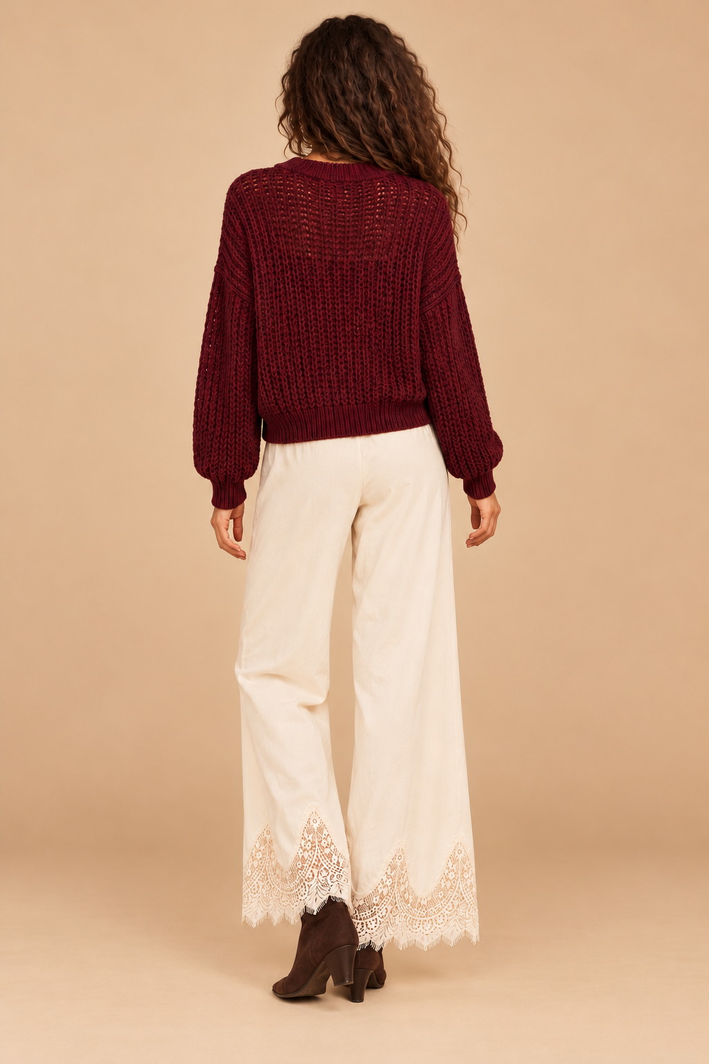 Julia Knit Sweater in Wine — back view on holiday model, cream lace pants and chocolate boots. Light tan studio background.