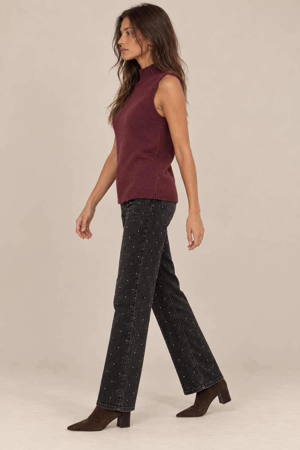 Sleeveless Knit Mock Neck Top in Wine — side view on holiday model, rhinestone black jeans and chocolate boots. Light tan studio background.