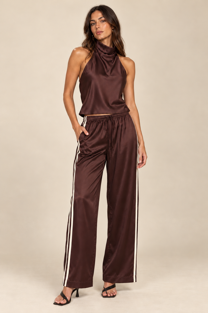 Brown Natural satin track pants and brown Romy halter — front view with natural pocket hand in front of cream side stripes, holiday model on light tan studio background.